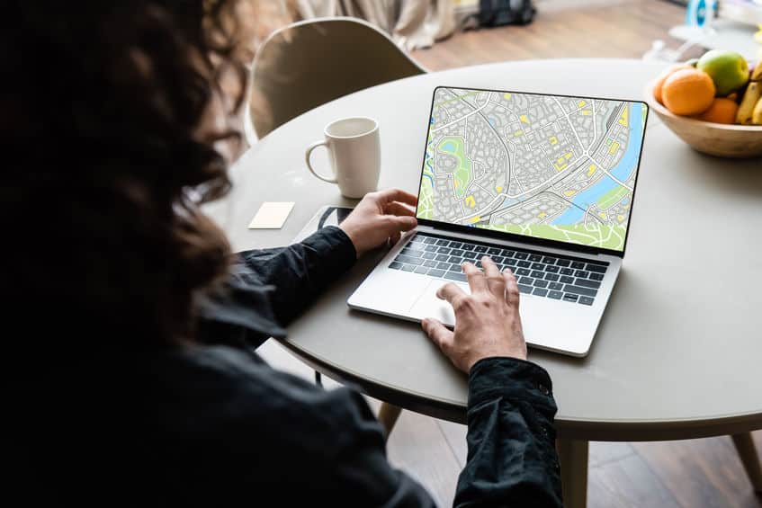 back view of freelancer using laptop with map on screen near coffee cup, sticky notes and fruits